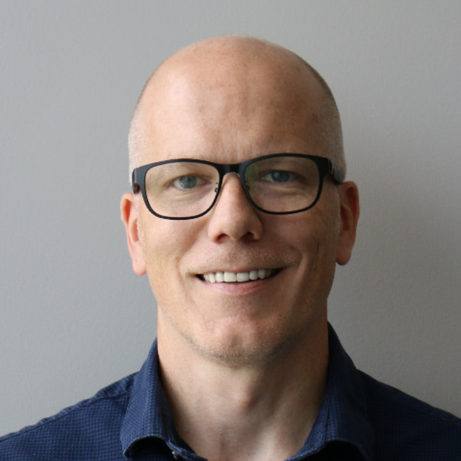 a smiling man wearing glasses in front of a grey background