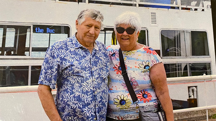 A couple stands together in front of a white boat with windows and a railing visible behind them